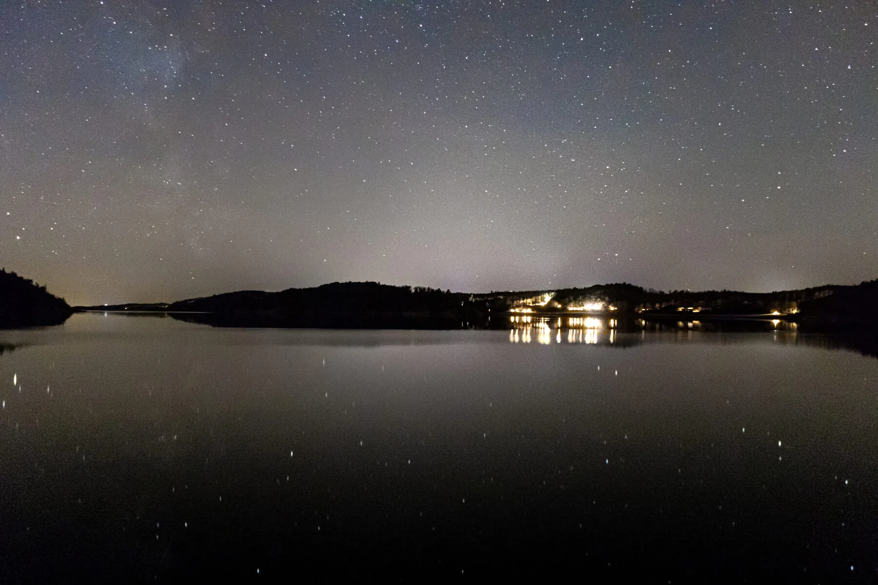 Sternenhimmel über einem ruhigen Gewässer, am Horizont spiegeln sich warme Ortslichter im Wasser