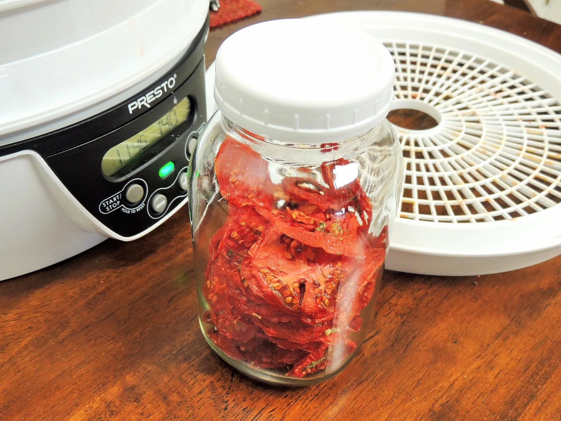 Ein verschlossenes Schraubglas mit getrockneten Tomaten steht neben einem Dörrautomaten auf einem Holztisch
