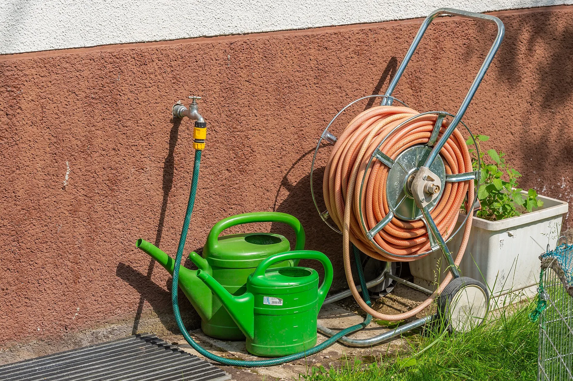 Wasserhahn an der Hauswand mit gelber Klickkupplung, zwei grüne Gießkannen und ein Schlauchwagen mit aufgerolltem Gartenschlauch