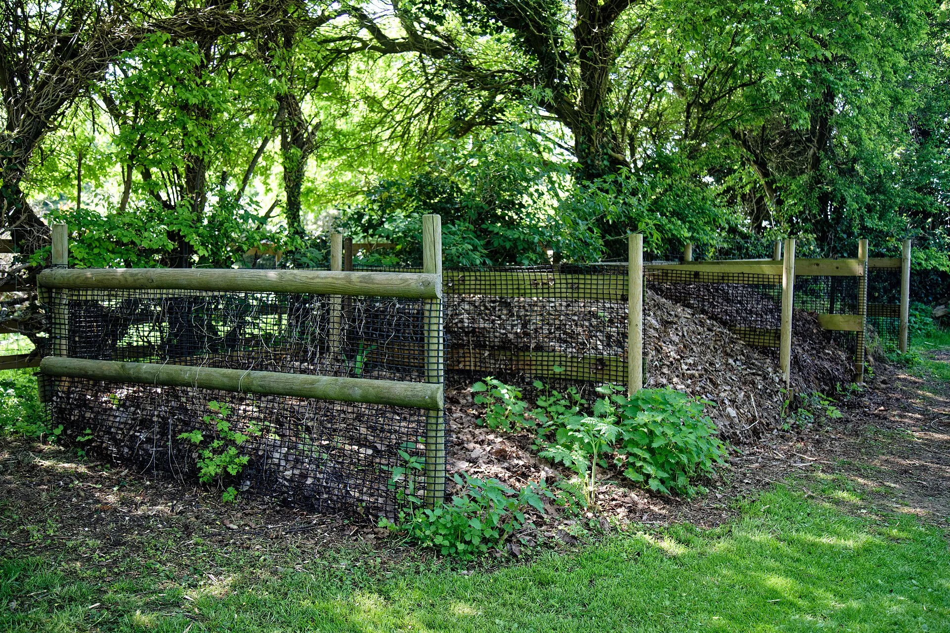 Offener Komposthaufen im Garten mit Holzpfosten und Drahtgitter, gefüllt mit Laub und Gartenabfällen, daneben Brennnesseln
