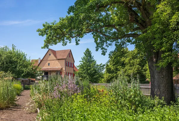 Bauerngarten mit sandigem Weg, Staketenzaun und blühenden Beeten vor einem alten Bauernhaus