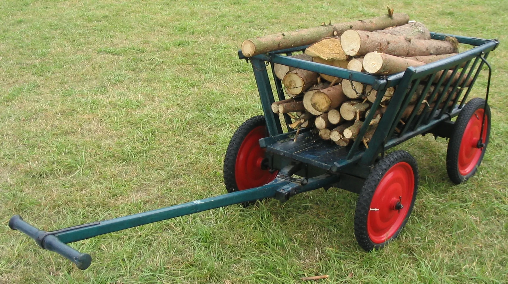 Hölzerner Bollerwagen mit vier Rädern und Zugstange, beladen mit Brennholzstücken, auf einer Wiese