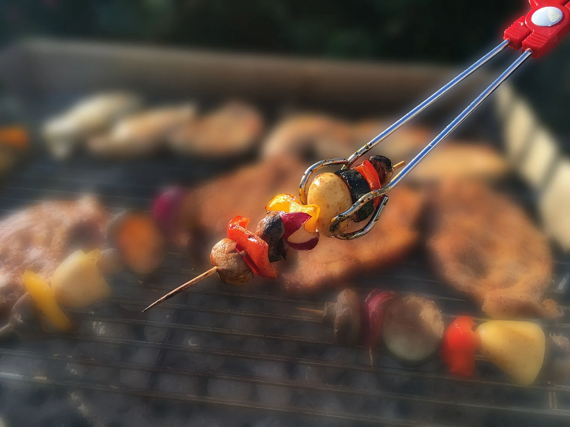 Vegetable skewer with bell pepper, potato, zucchini and mushroom is turned over the coals with a grill tongs