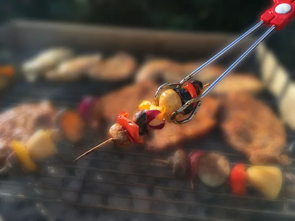 Vegetable skewer with bell pepper, potato, zucchini and mushroom is turned over the coals with a grill tongs