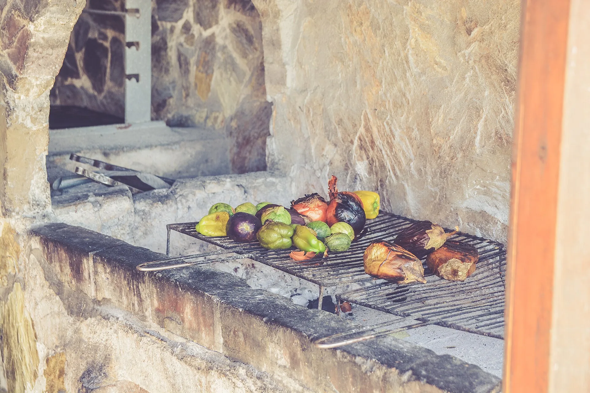 Peppers, aubergines and onions lying on the grate of a masonry fire pit over pale embers